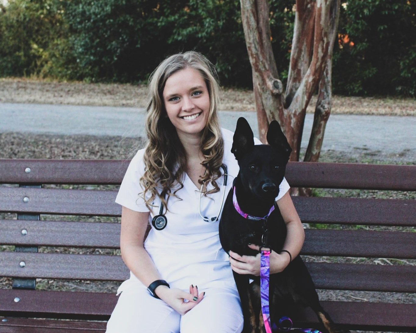 Smiling woman in a white medical uniform sitting on a bench with a black dog, showcasing a friendly bond in a natural outdoor setting.
