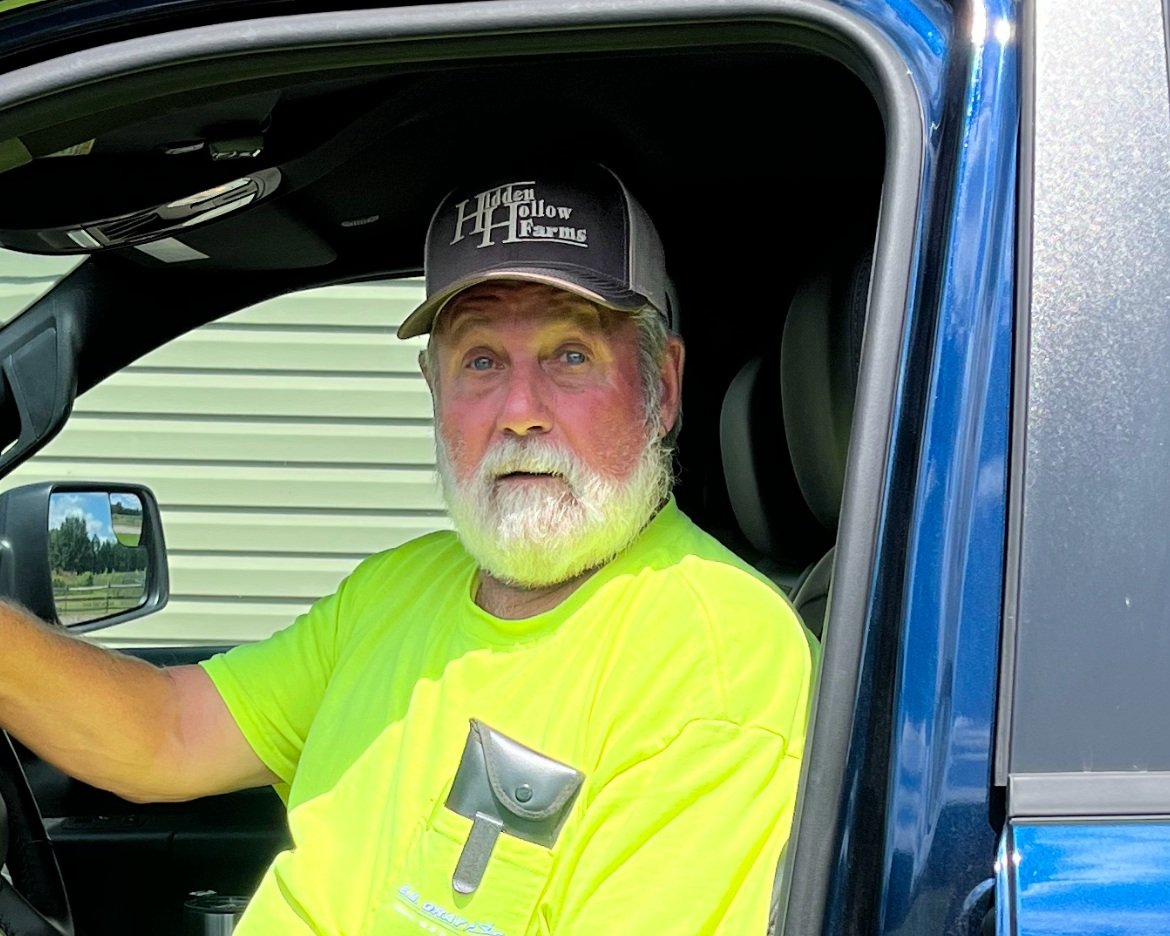 Man with a beard wearing a bright yellow shirt and a gray cap sits in the driver's seat of a blue truck, looking directly at the camera. The background features a green building and a clear sky, suggesting an outdoor farming or rural setting.