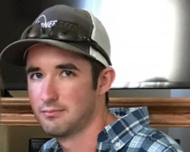 Young man wearing a gray baseball cap and sunglasses, with a plaid shirt, sitting indoors near a wooden railing.