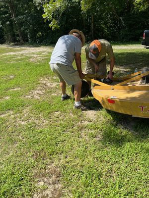 Two men working together to lift and secure a yellow boat trailer on grassy terrain, surrounded by trees and a truck in the background.
