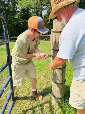 Two men working together to secure a fence post in a grassy field, one using a tool while the other assists, showcasing teamwork in outdoor maintenance.